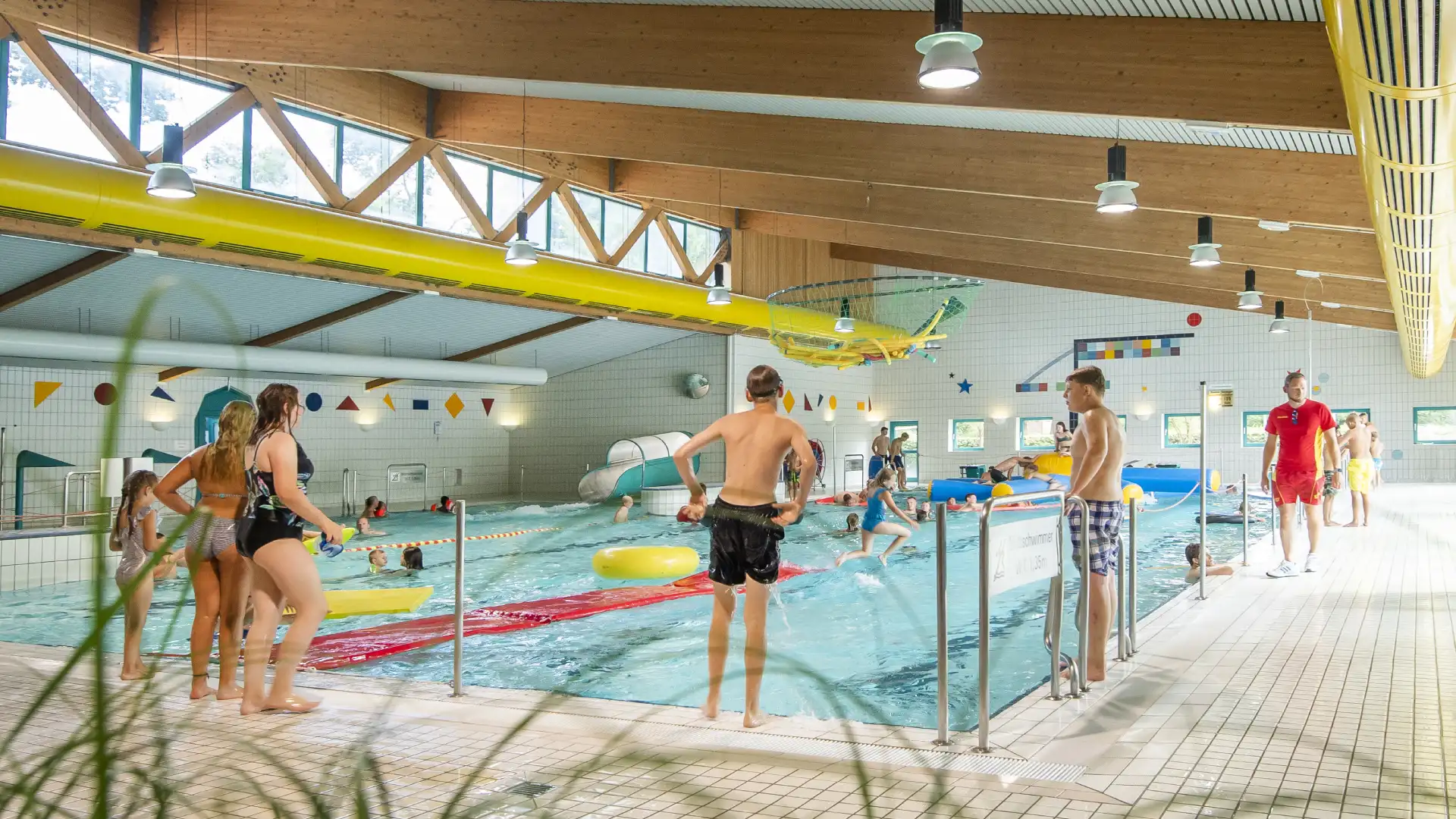 Ein Schwimmbad mit Kindern und Erwachsenen, die im Wasser spielen, während ein Rettungsschwimmer Aufsicht führt.